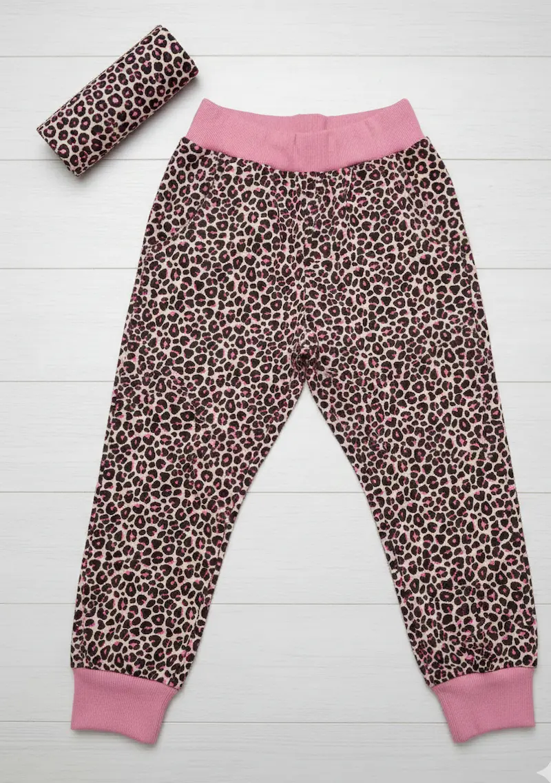 Produktansicht eines French Terry Stoffes als Meterware mit Leopard-Muster in Pink und Schwarz. Das Bild zeigt eine daraus genähte Kinder-Jogginghose mit rosa Bündchen sowie eine dekorative Stoffrolle auf einem weißen Holzhintergrund.