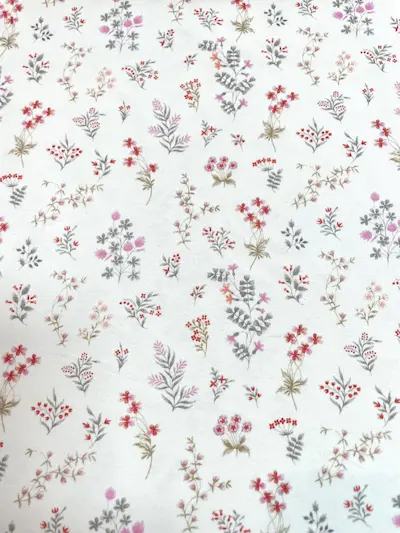 Weißer Jersey-Stoff mit einem feinen, floralen Muster aus kleinen bunten Wiesenblumen in Rosa, Rot und Grau.
