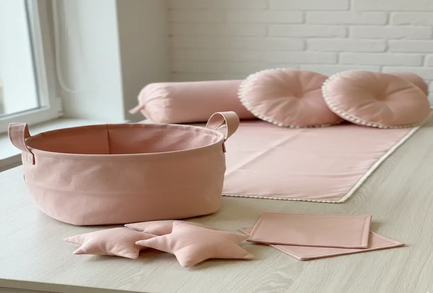 Hell gestalteter Raum mit handgemachten Wohnaccessoires aus unifarbenem hellrosafarbenem Baumwollstoff, bestehend aus einem Aufbewahrungskorb mit Henkeln, Sternenkissen, runden Dekokissen mit Pompon-Borte und einer passenden Nackenrolle auf einer hellen H
