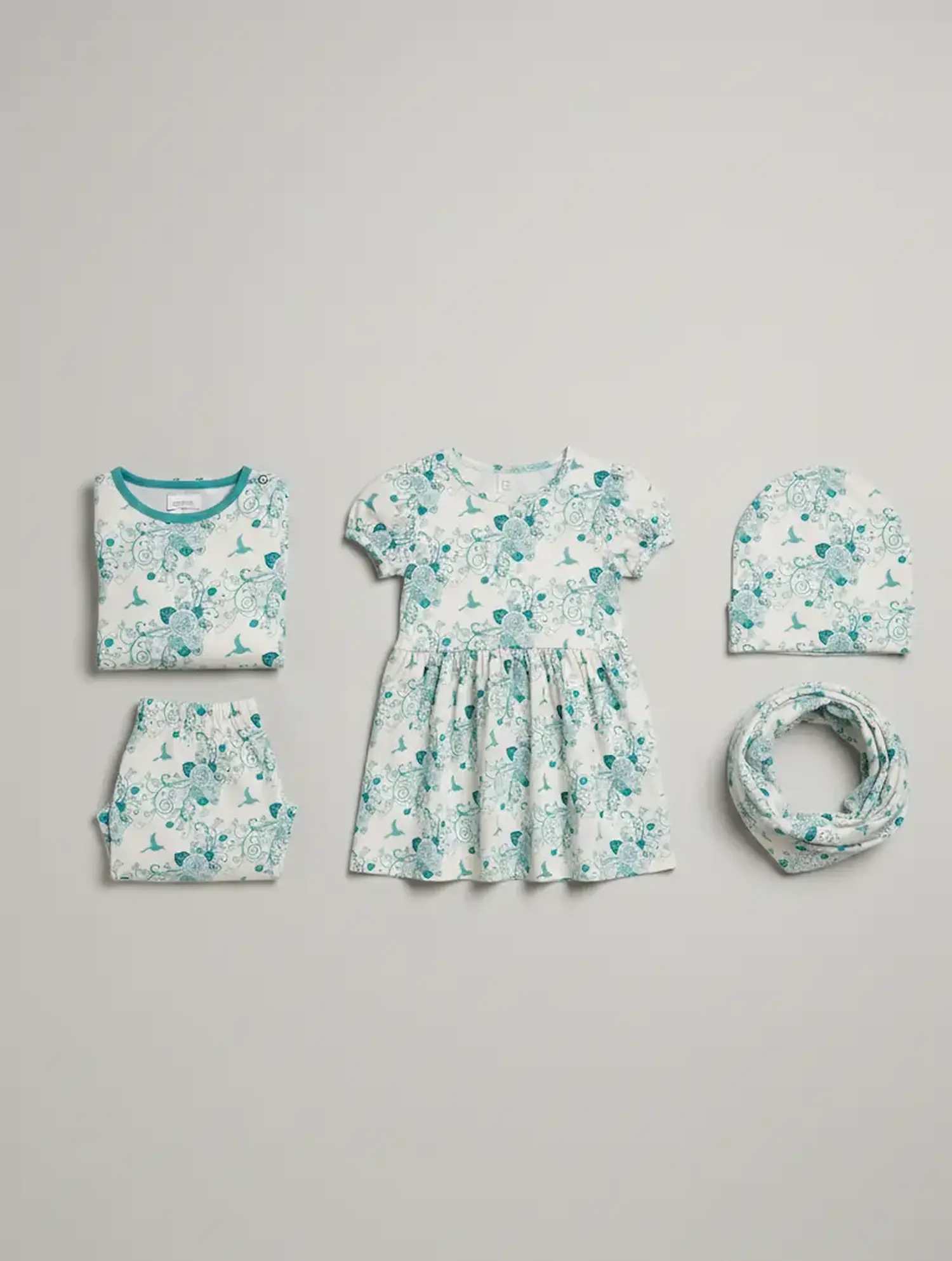 Eine Kollektion selbstgenähter Baby- und Kinderkleidung liegt ordentlich arrangiert auf einem hellgrauen Hintergrund. Alle Teile – darunter ein kurzärmeliges Kleidchen, ein gefaltetes T-Shirt, eine kleine Hose, eine Beanie-Mütze und ein Loop-Schal – sind