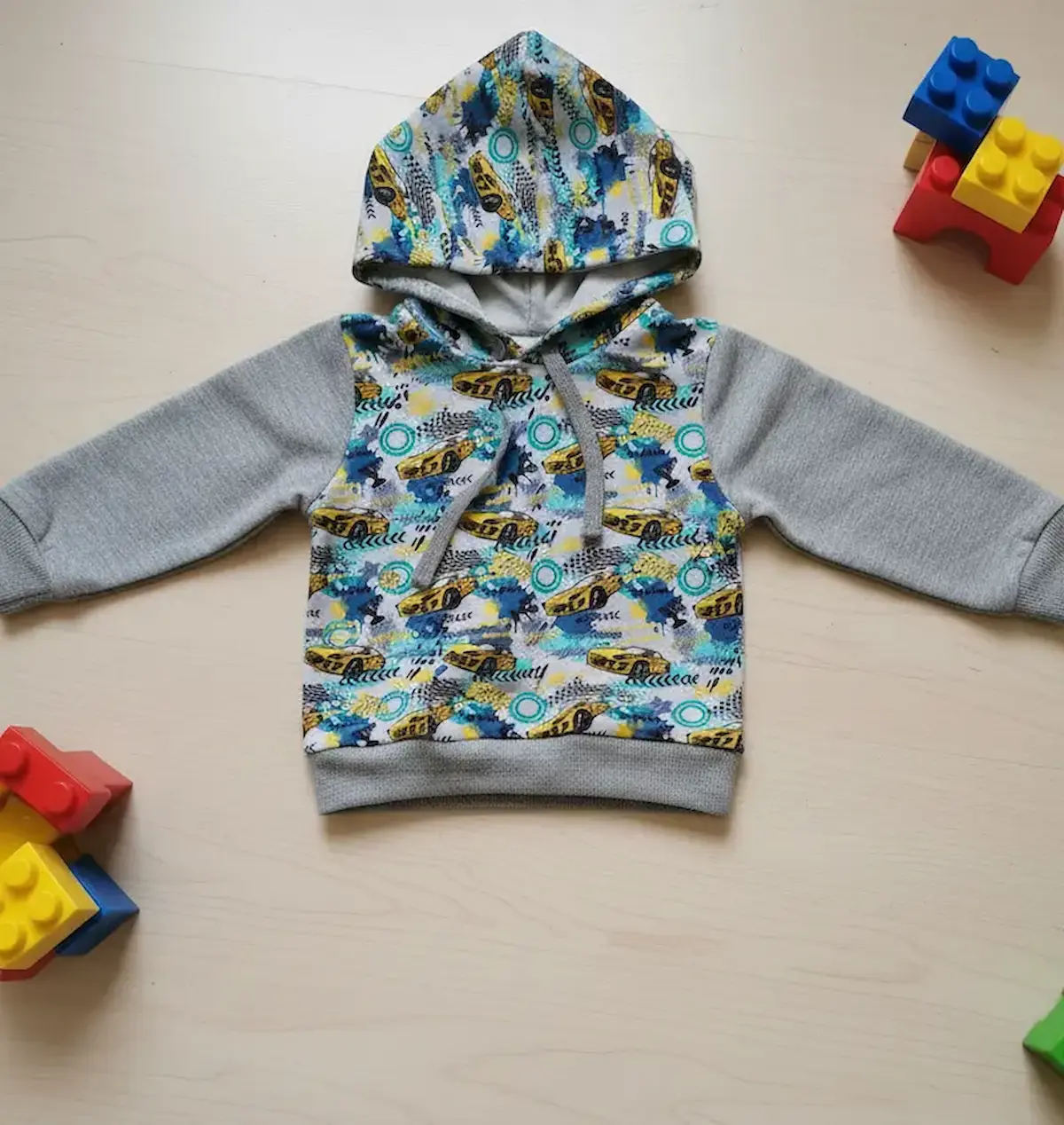 Cooles Flatlay-Nähbeispiel: Ein selbstgenähter Kinder-Hoodie aus French Terry mit gelben Rennwagen und Graffiti-Muster, kombiniert mit grauen Ärmeln auf hellem Holzboden mit Spielzeugsteinen.
