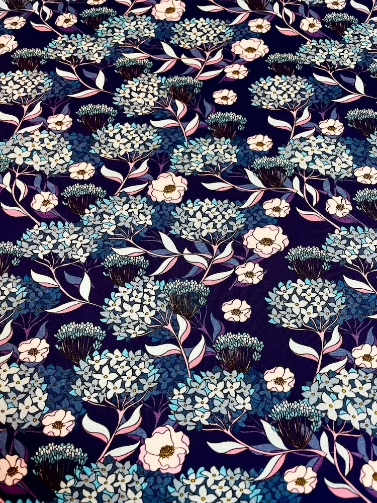 Detailgetreue Draufsicht eines Jersey-Stoffs mit Hortensien-Motiv. Das Muster zeigt stilisierte hellblaue und beerenfarbene Blüten auf dunkelblauem Grund. Klare Darstellung des floralen Drucks für Nähprojekte.