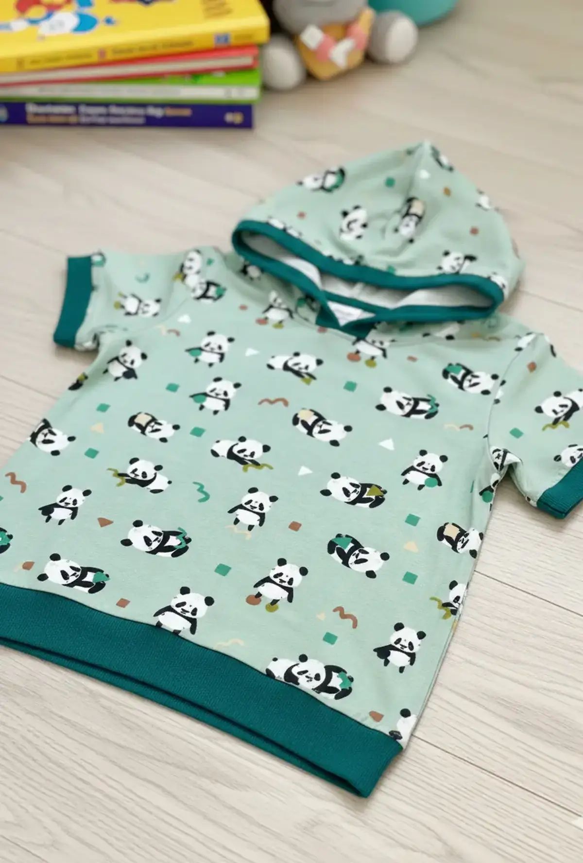 Genähtes Kurzarm-Kapuzenshirt für Kinder aus salbeigrünem Panda-Baumwolljersey, kombiniert mit kontrastierenden Bündchen und Kapuzeneinfassung in Smaragdgrün.
