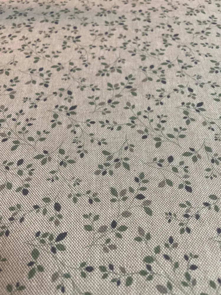 Strukturierter Dekostoff als Meterware mit einem feinen Streumuster aus kleinen grünen Blättern und Zweigen auf beigem Naturhintergrund.