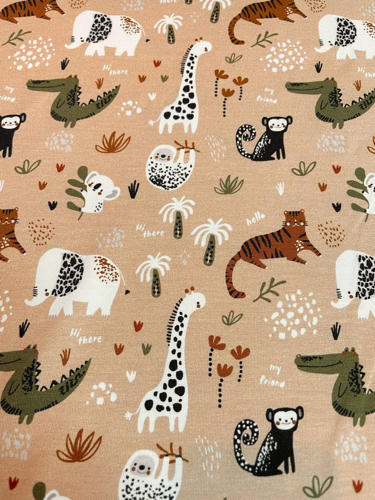 Ein flach liegender Baumwolljersey-Stoff als Meterware in hellem Apricot-Braun, bedruckt mit einem bunten Kindermuster aus Dschungeltieren wie Giraffen, Elefanten, Tigern, Krokodilen, Faultieren und kleinen Palmen.