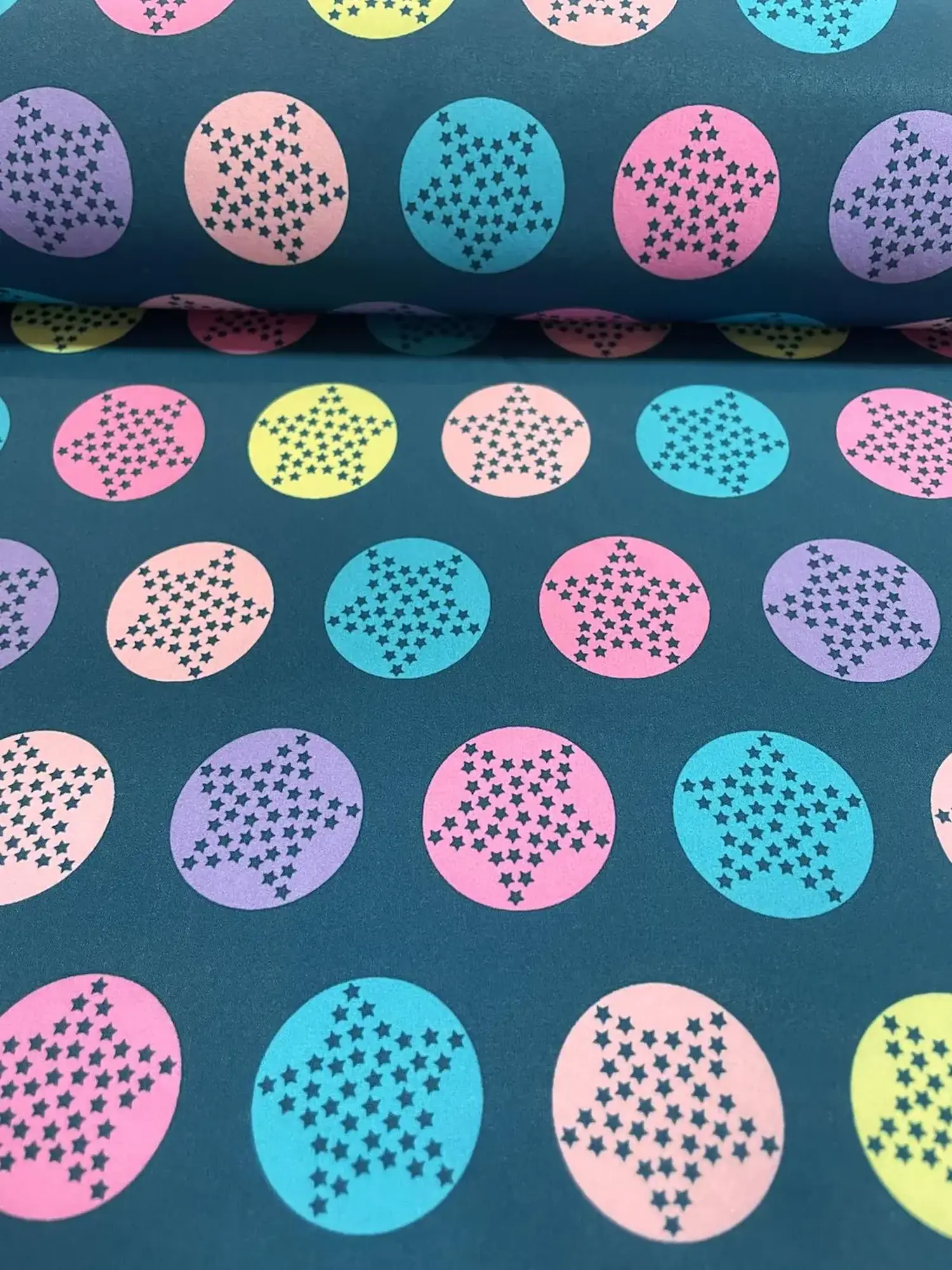 Detailaufnahme eines hochwertigen Softshell-Stoffs in Dunkelblau. Das Muster zeigt farbige Kreise in Neon-Pink, Türkis, Flieder und Hellgelb, in denen durch kleine Aussparungen große Sternenmotive entstehen. Funktionaler Outdoorstoff für Kinderbekleidung.