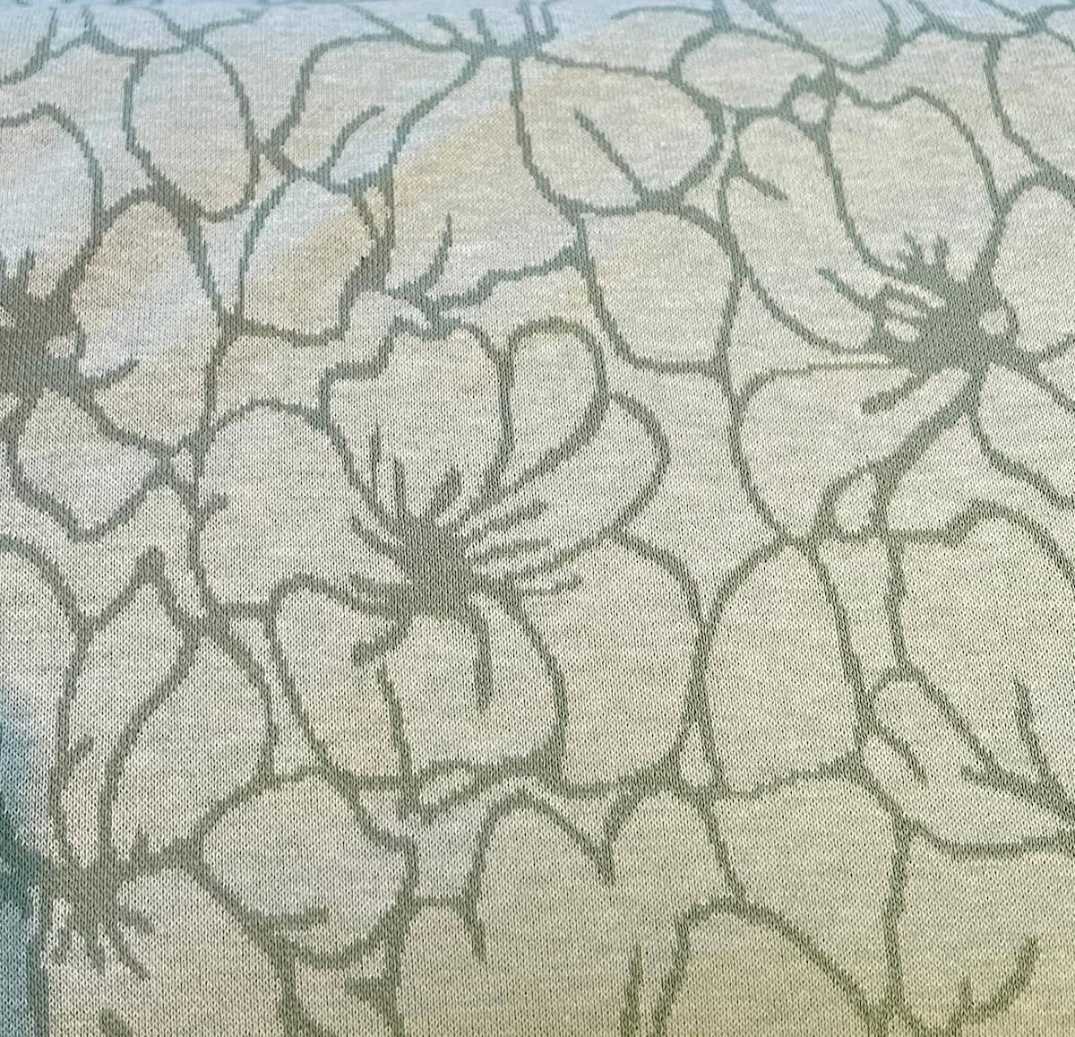 Hochwertiger Strickjacquard-Stoff mit großflächigem Blumenmuster. Die Konturen der Blüten sind in Dunkelgrün auf einem melierten Hintergrund in Hellgrün und Beige gestrickt.