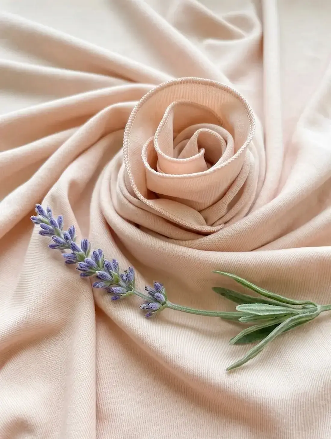 Nahaufnahme von pudderrosa Baumwolljersey, zu einer Rose drapiert, dekoriert mit einem frischen Lavendelzweig.
