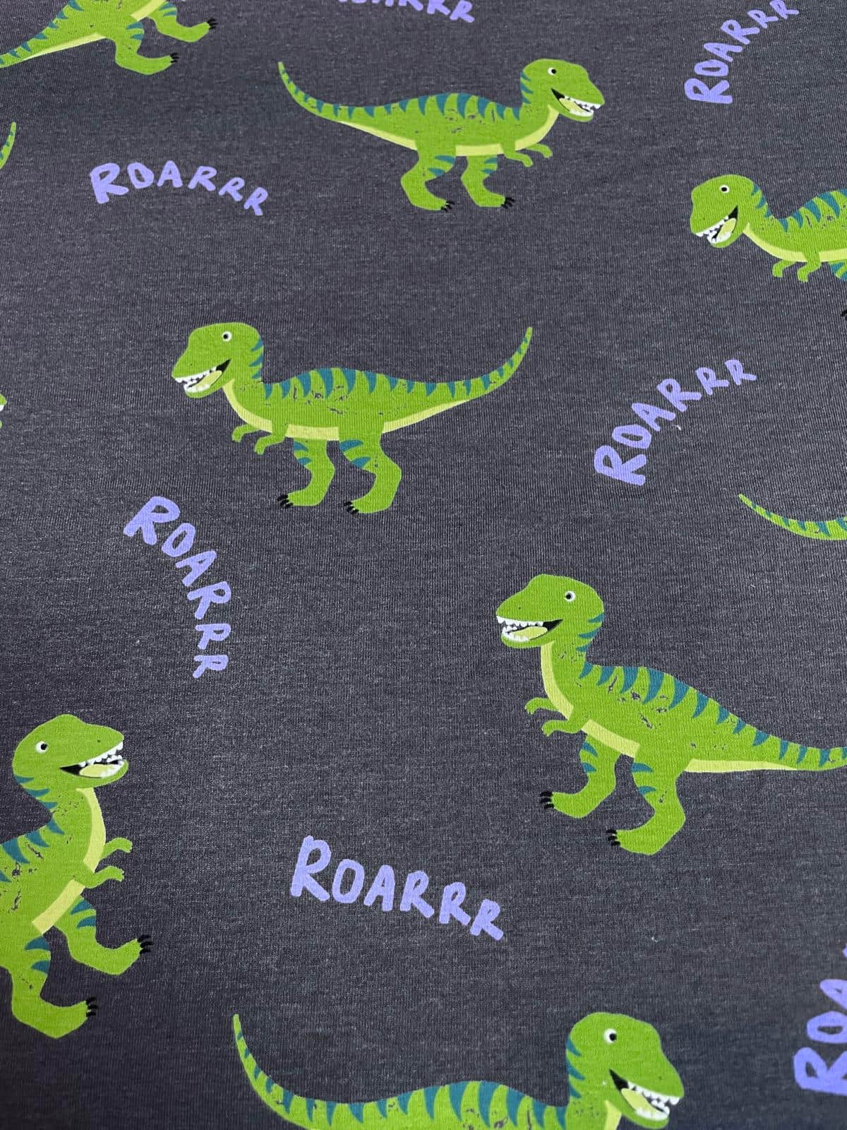 Ein flach liegender Baumwolljersey-Stoff als Meterware in Anthrazit / Dunkelgrau, bedruckt mit einem coolen Kindermuster aus grünen, gestreiften Dinosauriern (T-Rex) und lilafarbenen „ROARRR“-Schriftzügen.