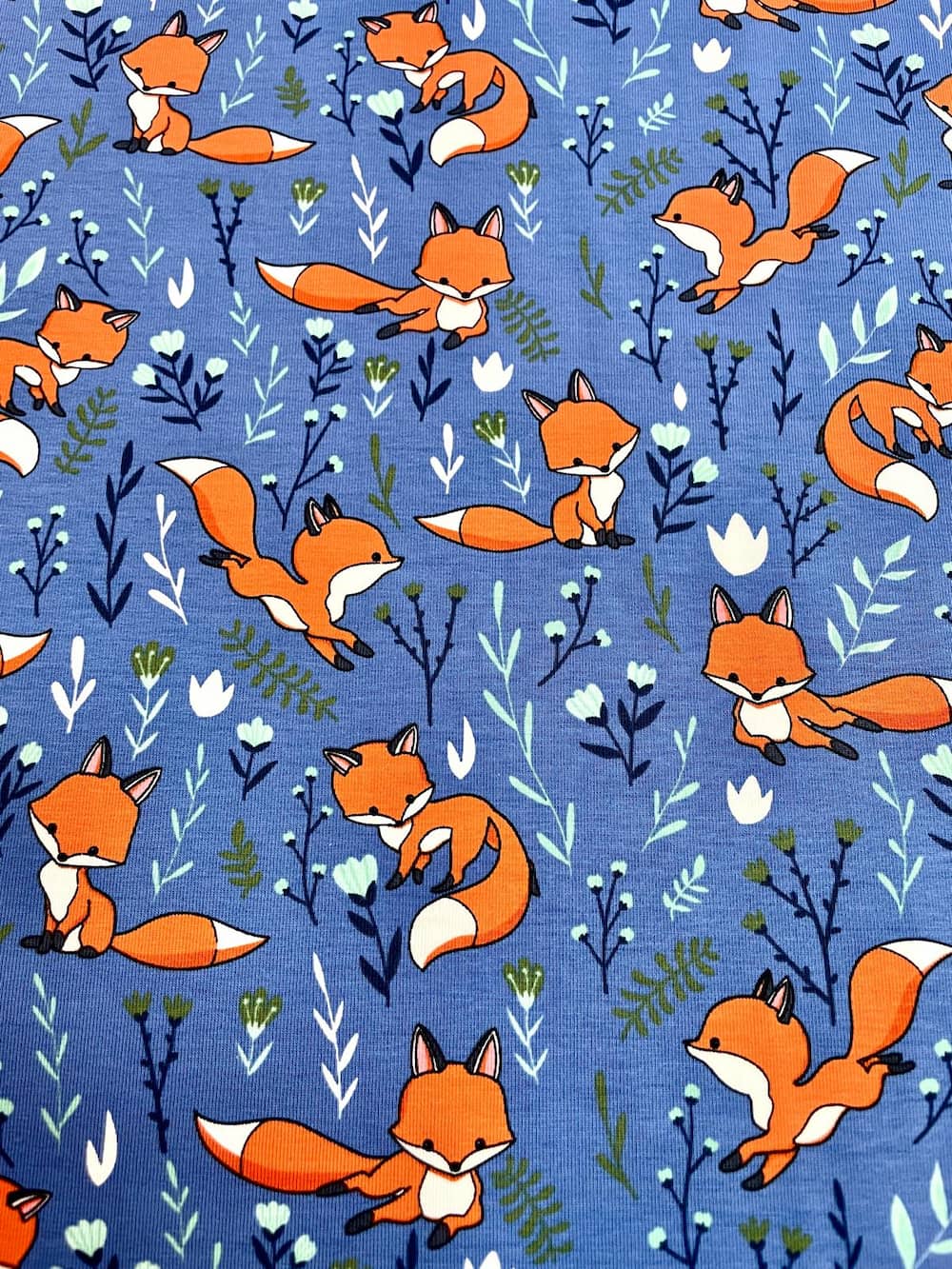 Hochwertiger Baumwolljersey in kräftigem Blau mit verschiedenen Fuchs-Motiven und kleinen floralen Elementen wie Farnen und Blüten, ideal für Kinder-Shirts und Babykleidung.