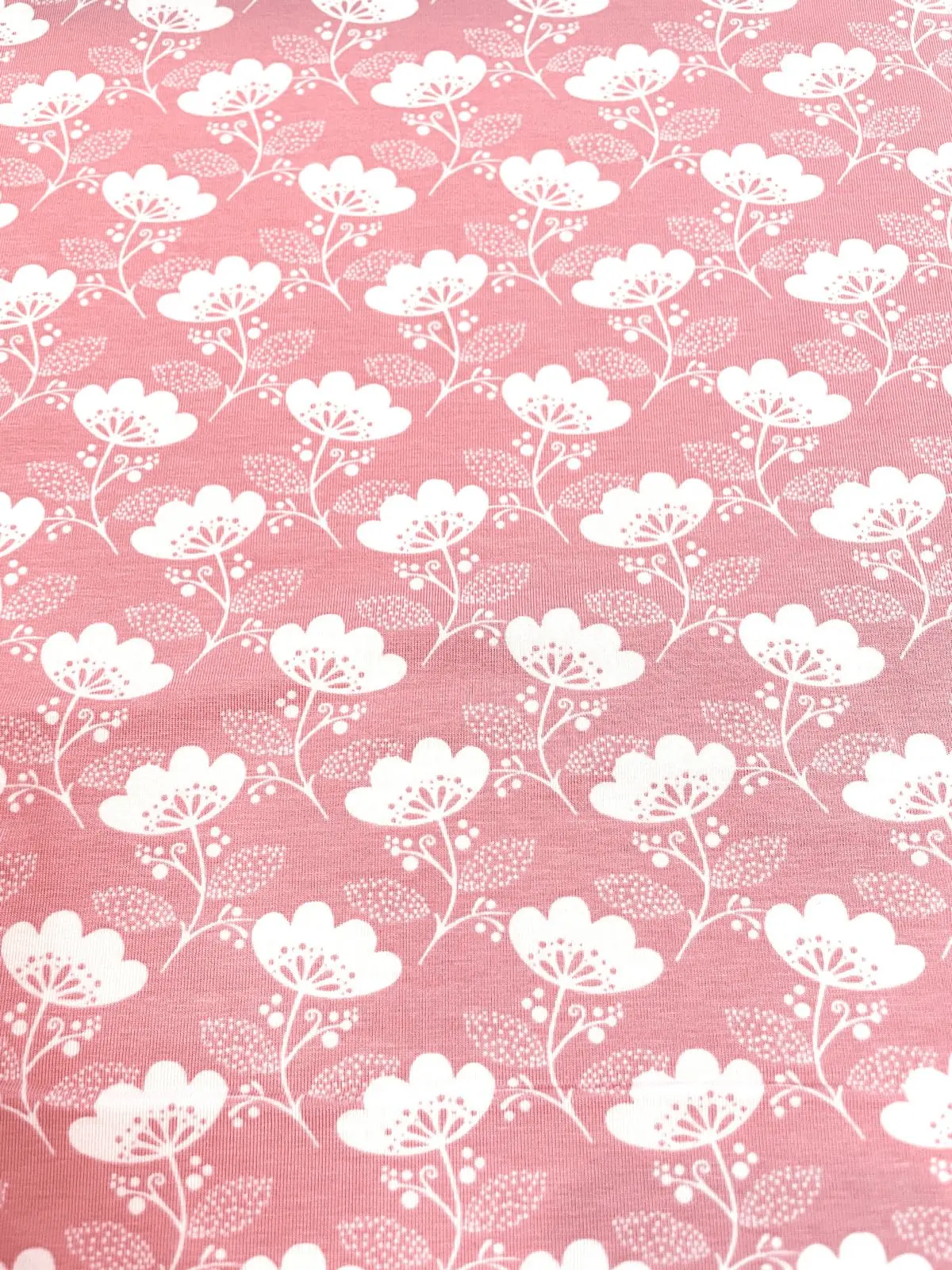 Ein flach liegender Baumwolljersey-Stoff als Meterware in einem hellen Altrosa / Rosa, bedruckt mit einem eleganten, gleichmäßigen Muster aus stilisierten weißen Blumen, feinen Blättern und kleinen weißen Beerenstrukturen.