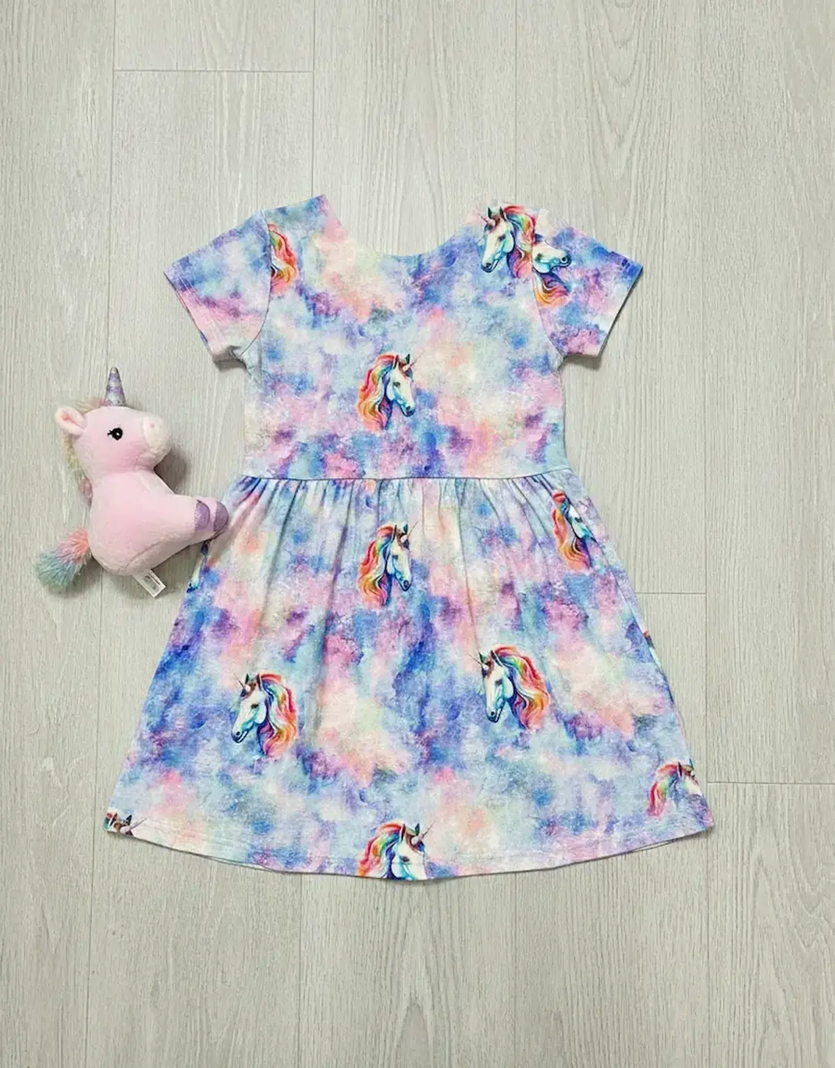 Ein selbstgenähtes Mädchenkleid aus buntem Baumwolljersey (Meterware) mit regenbogenfarbenem Wolkenmuster und Einhorn-Motiven, flach liegend auf Holzboden neben einem kleinen Plüscheinhorn.