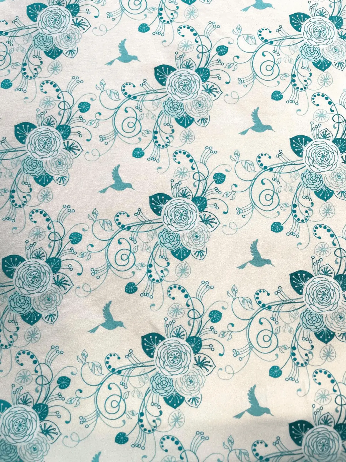 Hochwertiger, elastischer Baumwolljersey als Meterware mit weißem Hintergrund. Der Stoff ist mit einem filigranen, floralen Muster in verschiedenen Türkis- und Minttönen bedruckt, das fliegende Kolibris, kunstvolle Rosenblüten und verschnörkelte Blattrank