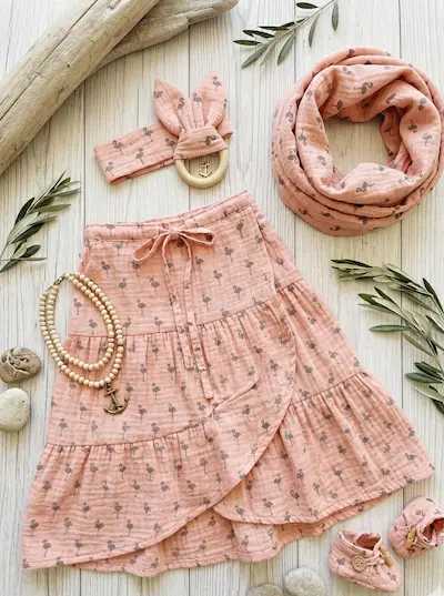 Musselin-Stoff in Apricot mit grauem Flamingo-Muster, verarbeitet zu einem Kinder-Set mit Stufenrock, Loop-Schal, Stirnband und Babyschuhen auf hellem Holzboden.