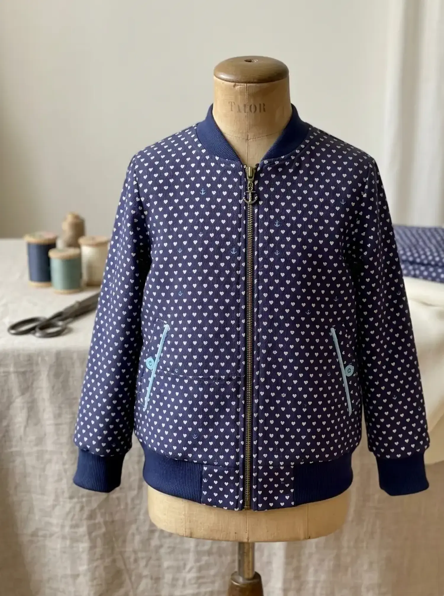 Blaue Bomberjacke für Kinder aus Mors Crepe Baumwollstoff mit weißem Herzmuster und hellblauen Akzenten auf einer Schneiderpuppe