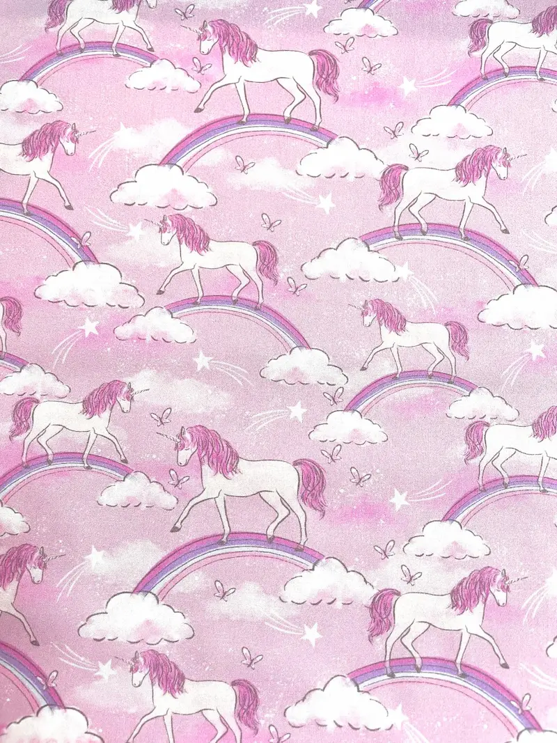 Draufsicht auf rosa Baumwoll-Webware als Meterware mit einem sich wiederholenden Motiv aus weißen Einhörnern, regenbogenfarbenen Bögen, weißen Wolken und kleinen Sternen.