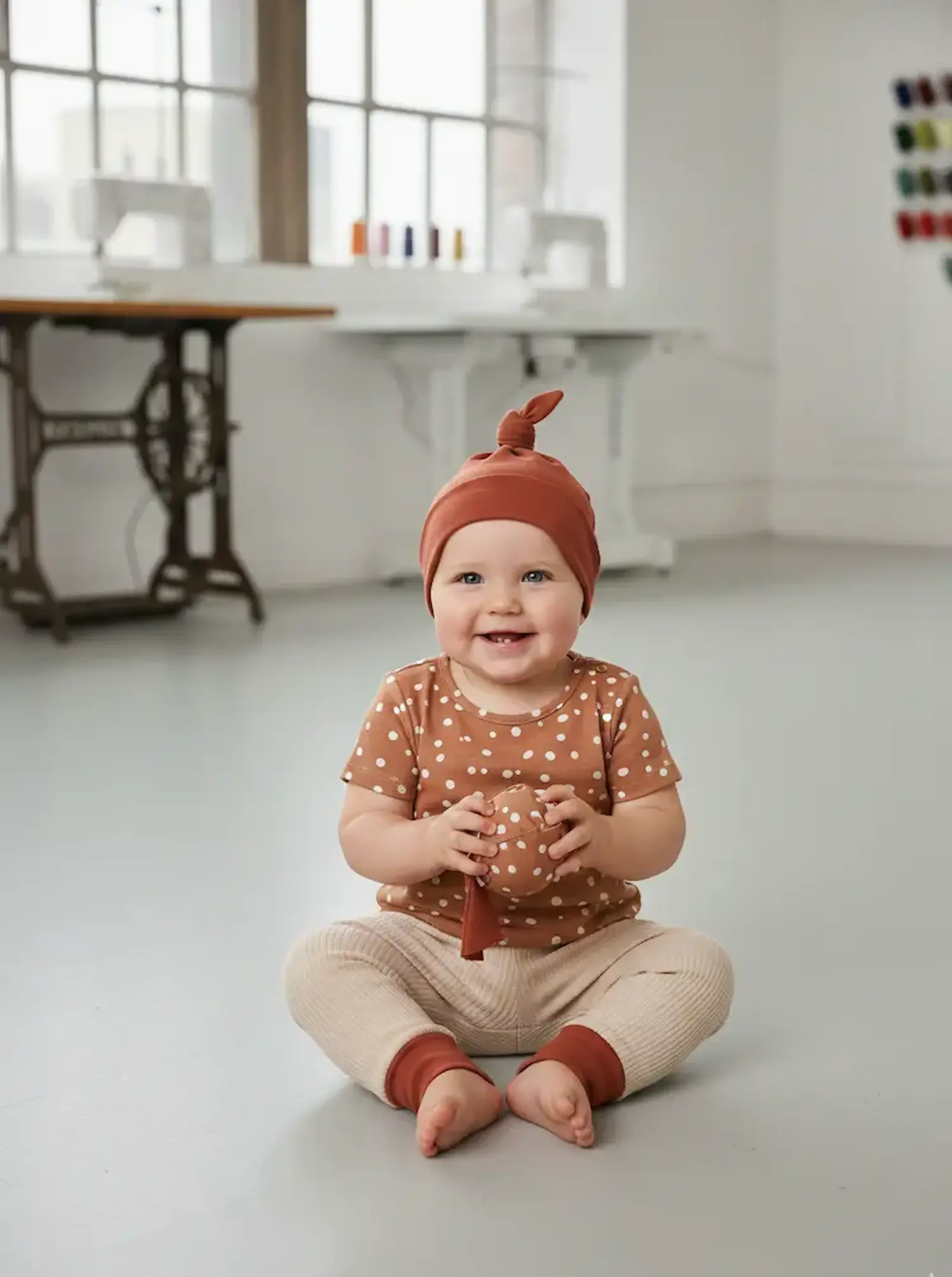 Ein sitzendes Baby in einem hellen Näh-Atelier trägt ein Kurzarm-Shirt aus altrosa Jerseystoff mit weißen Punkten, eine passende Knotenmütze und hält einen selbstgenähten Stoffball in den Händen.
