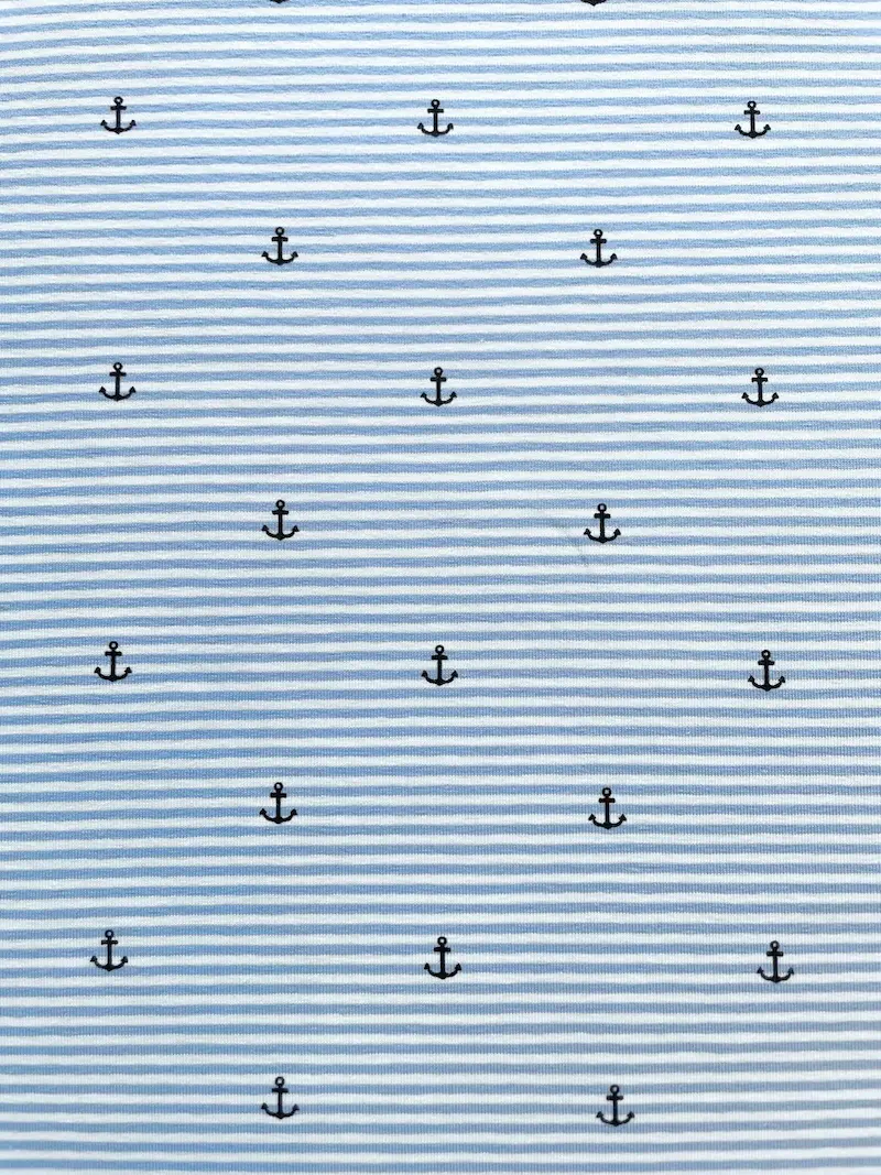 Nahaufnahme von elastischem Baumwolljersey in Hellblau und Weiß feingestreift, verziert mit einem regelmäßigen Muster aus kleinen dunkelblauen Ankern; hochwertige Meterware für maritime Babykleidung, T-Shirts und sommerliche Accessoires.