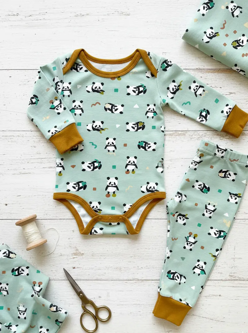 Flach liegendes, genähtes Baby-Set bestehend aus einem Langarmbody und einer passenden Hose aus salbeigrünem Panda-Baumwolljersey, eingefasst mit ockerbraunen Bündchen, dekoriert mit Schere und Garnrolle.