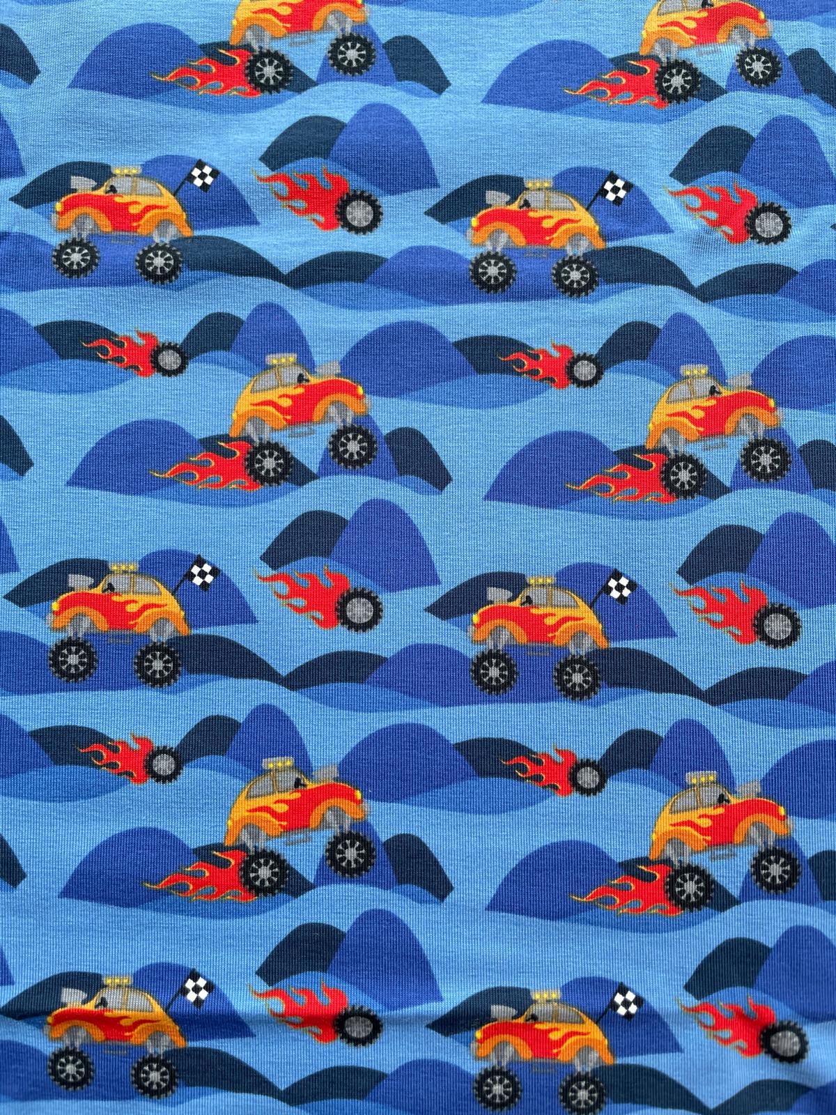 Blauer Baumwolljersey-Stoff mit Rapport-Muster aus roten und gelben Monster Trucks mit Flammen-Details auf dunkelblauen Hügeln.
