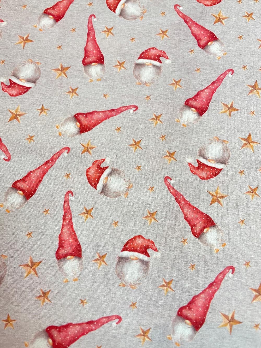 Hochwertiger Baumwollstoff als Meterware mit niedlichen Weihnachtswichteln mit roten Mützen und goldenen Sternen auf hellem grauem Hintergrund.