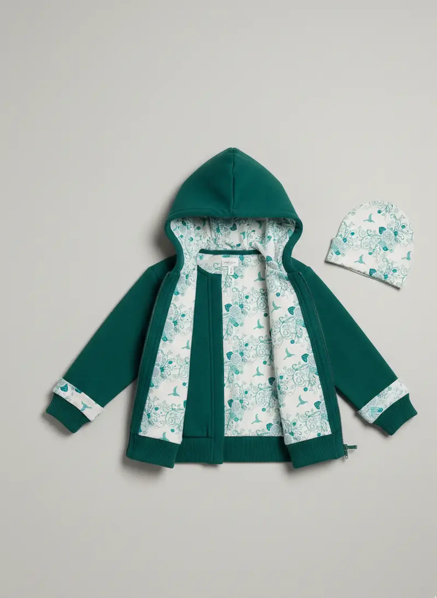 Eine petrolgrüne Kinder-Sweatjacke mit Kapuze liegt flach ausgebreitet vor einem grauen Hintergrund. Die Jacke ist innen komplett mit dem weißen Baumwolljersey gefüttert, der ein türkisfarbenes Muster aus Vögeln und Blumen zeigt. Rechts daneben liegt eine