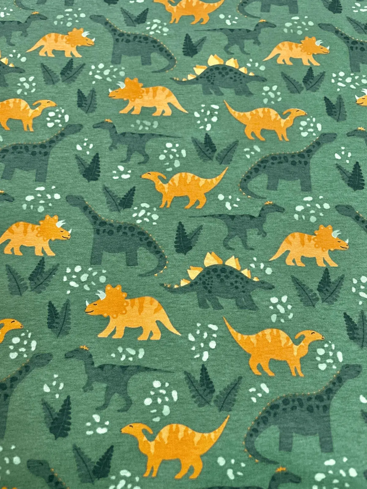 Grüner Baumwolljersey als Meterware, bedruckt mit einem dichten Muster aus orangefarbenen und dunkelgrünen Dinosauriern wie Triceratops und Stegosaurus zwischen Farnblättern.