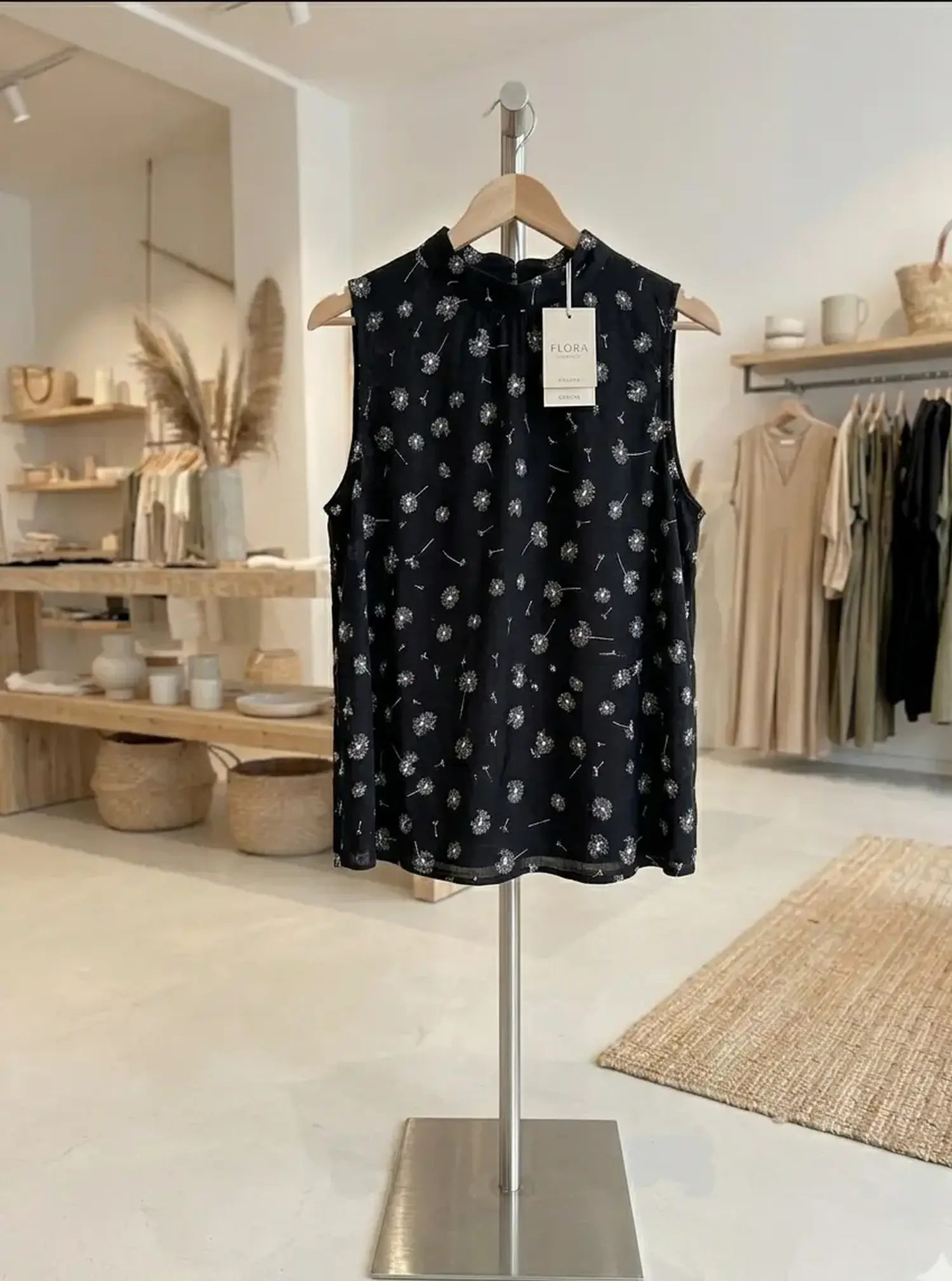 Ärmelloses Top aus schwarzer Viskose-Mix Webware mit weißem Pusteblumen-Muster auf einem Kleiderbügel in einer Boutique.