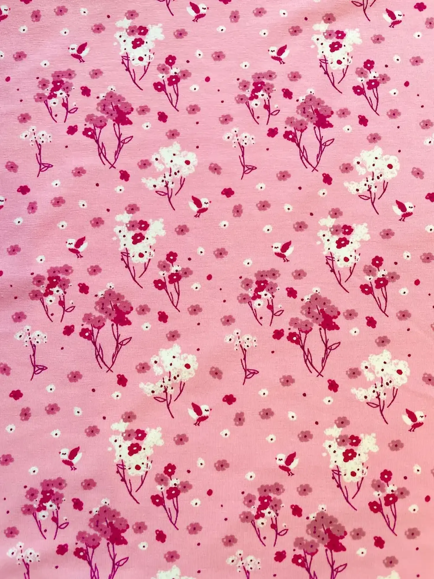Detaillierte Nahaufnahme von hellrosa Baumwolljersey als Meterware mit einem verspielten Allover-Print aus kleinen, fliegenden Vögeln in Pink und Weiß sowie zarten Frühlingsblumen und Blütenzweigen.