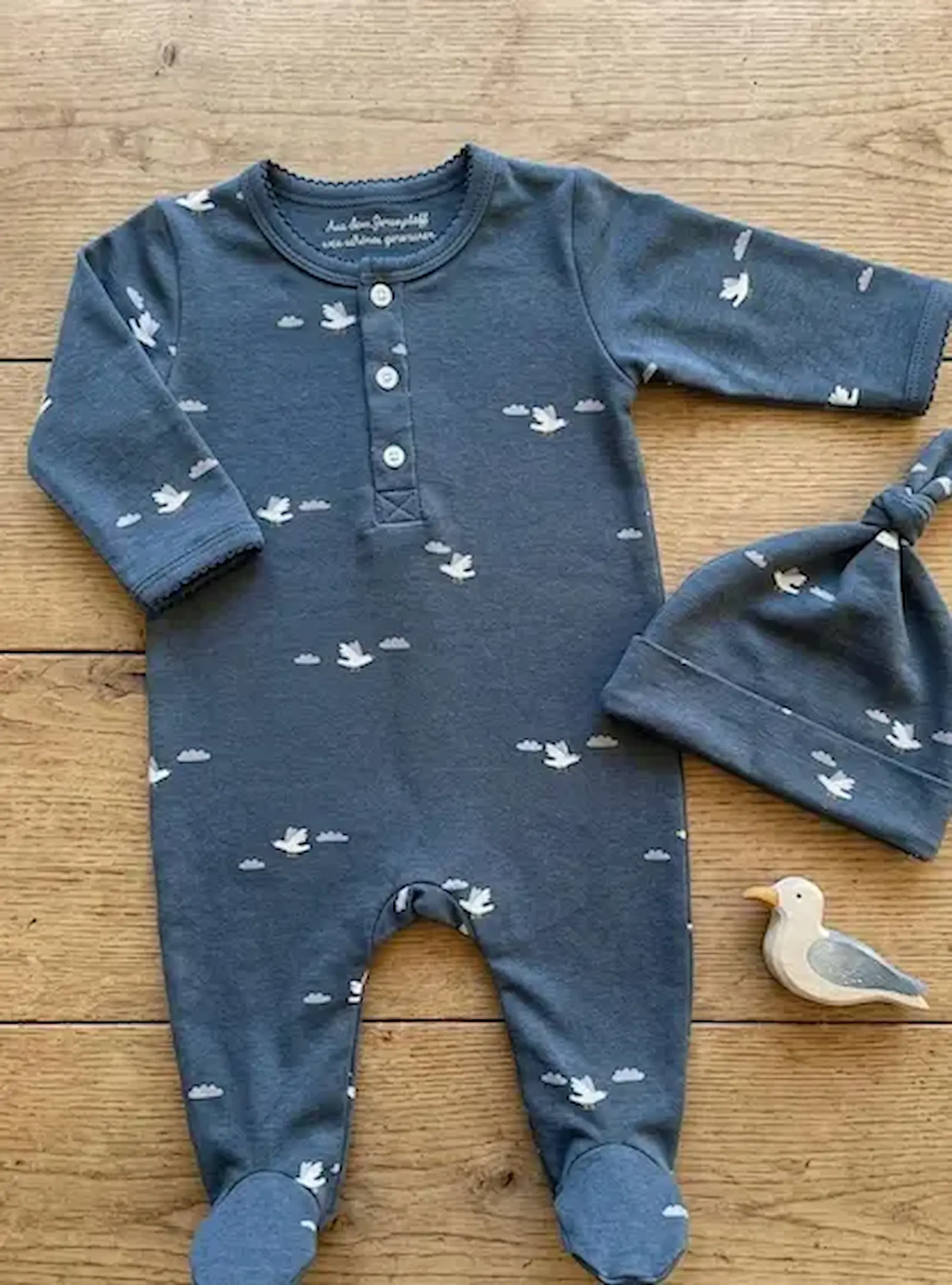 Ein blau melierter Baby-Schlafanzug mit passender Knotenmütze aus Jersey-Stoff, bedruckt mit weißen Möwen und kleinen Wolken, flach liegend auf Holzboden.