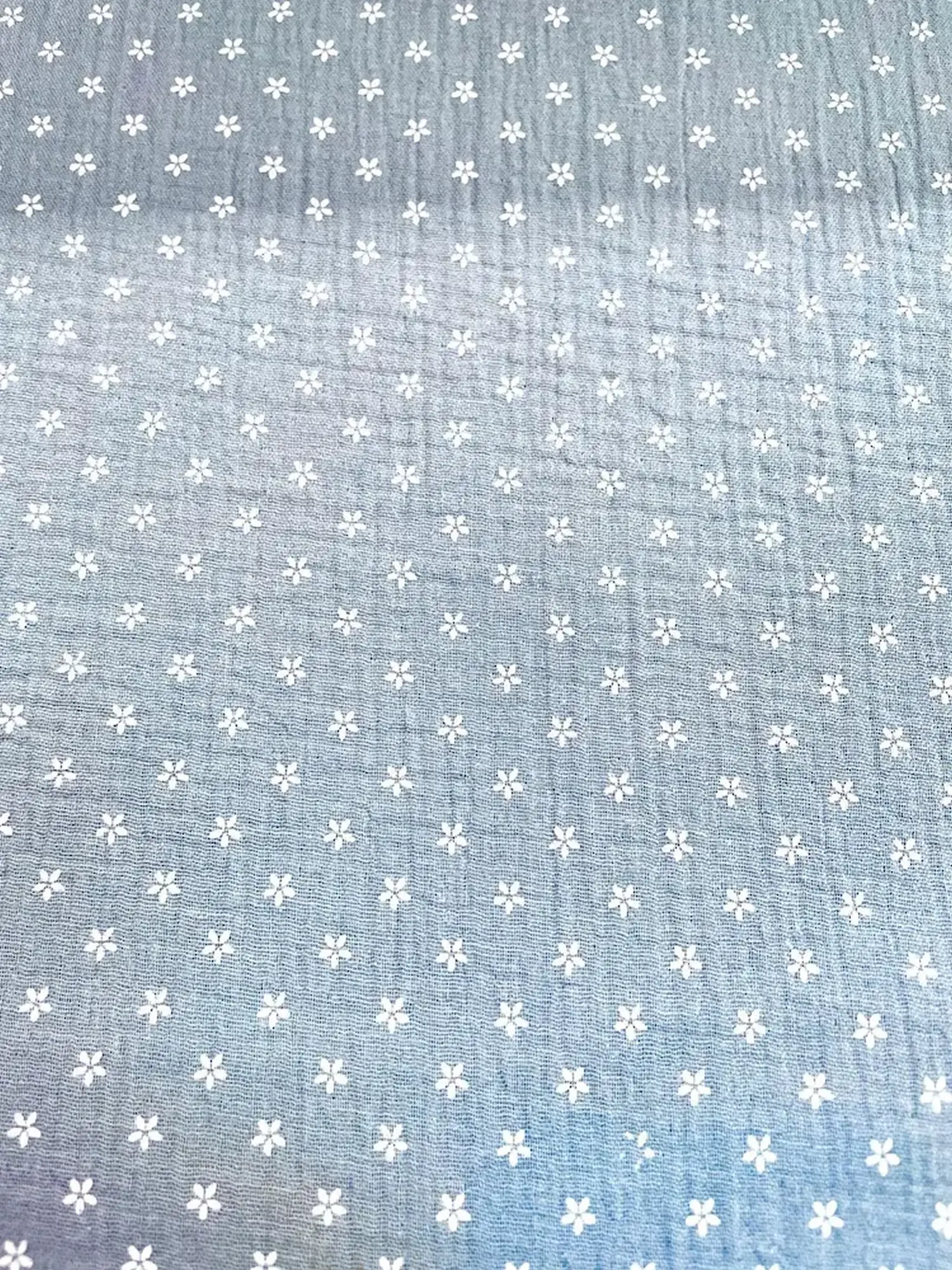 Flächenansicht von taubenblauem Musselin-Stoff mit einem Allover-Print aus kleinen weißen Blüten. Die Aufnahme zeigt die regelmäßige Musterverteilung und die charakteristische Crinkle-Struktur der Double-Gauze Meterware.
