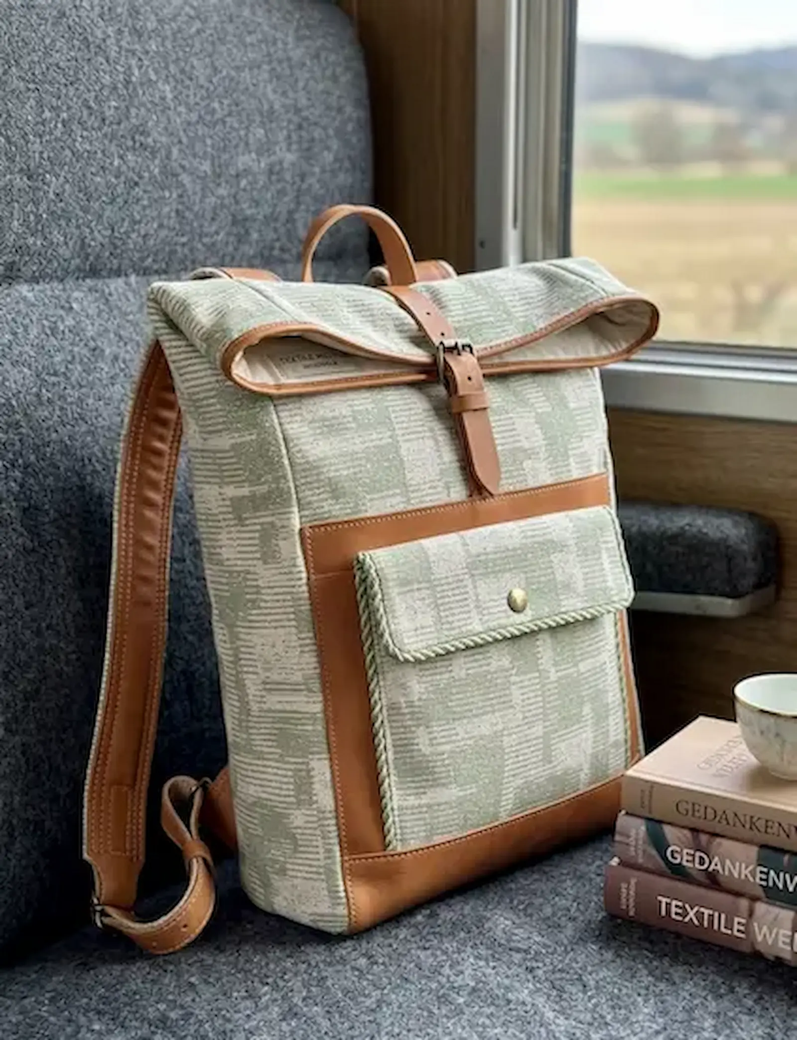 Moderner Roll-Top Rucksack aus grün-weißem Dekostoff mit abstrakter Webstruktur und braunen Leder-Details auf einem Zugsitz.
