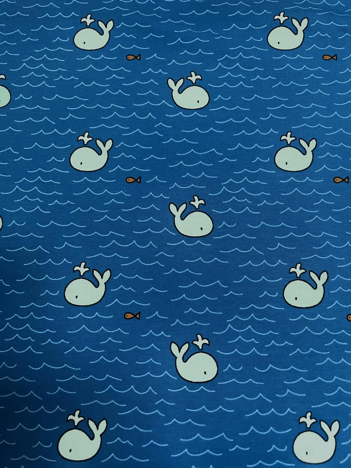 Ein flach liegender Baumwolljersey-Stoff als Meterware in hellem Dunkelblau, bedruckt mit einem maritimen Allover-Muster aus kleinen, hellblauen Walen mit Wasserfontänen, winzigen orangefarbenen Fischen und feinen, hellblauen Wellenlinien.