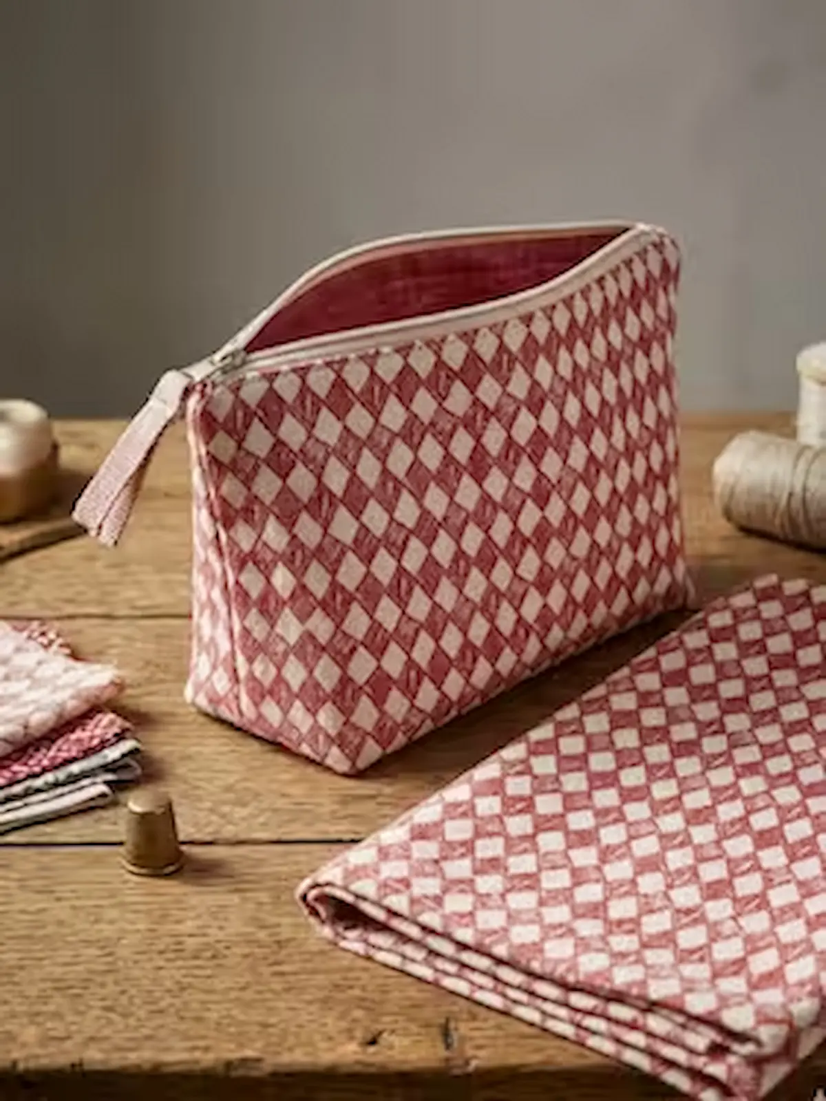 Eine genähte Kosmetiktasche mit Reißverschluss aus rotem Baumwoll-Popeline-Stoff mit weißem Rautenmuster steht auf einem Holztisch. Im Vordergrund liegt ein passend gefaltetes Stück der Meterware, im Hintergrund sind ein Fingerhut, Garnrollen und weitere