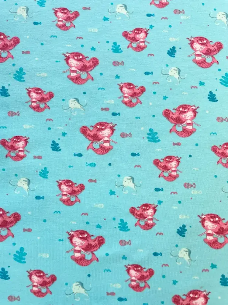 Nahaufnahme von hellblau-türkisfarbenem Jersey-Stoff als Meterware mit einem Allover-Print aus kleinen Meerjungfrauen mit pinken Haaren, kleinen hellen Quallen, Fischen und Seegras.