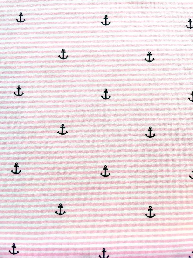 Nahaufnahme von elastischem Baumwolljersey in Rosa und Weiß gestreift, bedruckt mit kleinen dunkelblauen Ankern; hochwertige maritime Meterware für Kinderkleidung, sommerliche Kleider und Accessoires.