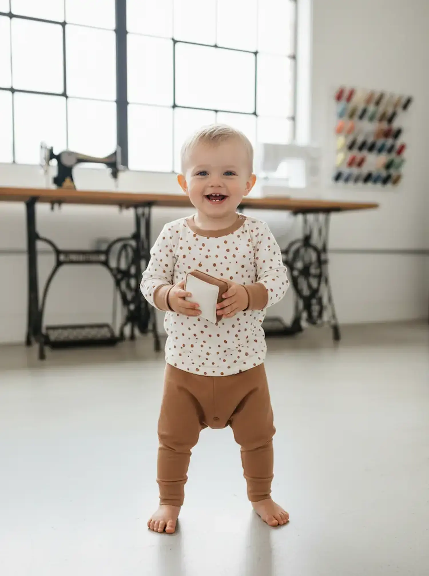 Ein lachendes Kleinkind steht in einem Näh-Atelier und trägt ein Langarmshirt aus cremeweißem Jerseystoff mit braunen Punkten, kombiniert mit einer passenden braunen Pumphose.