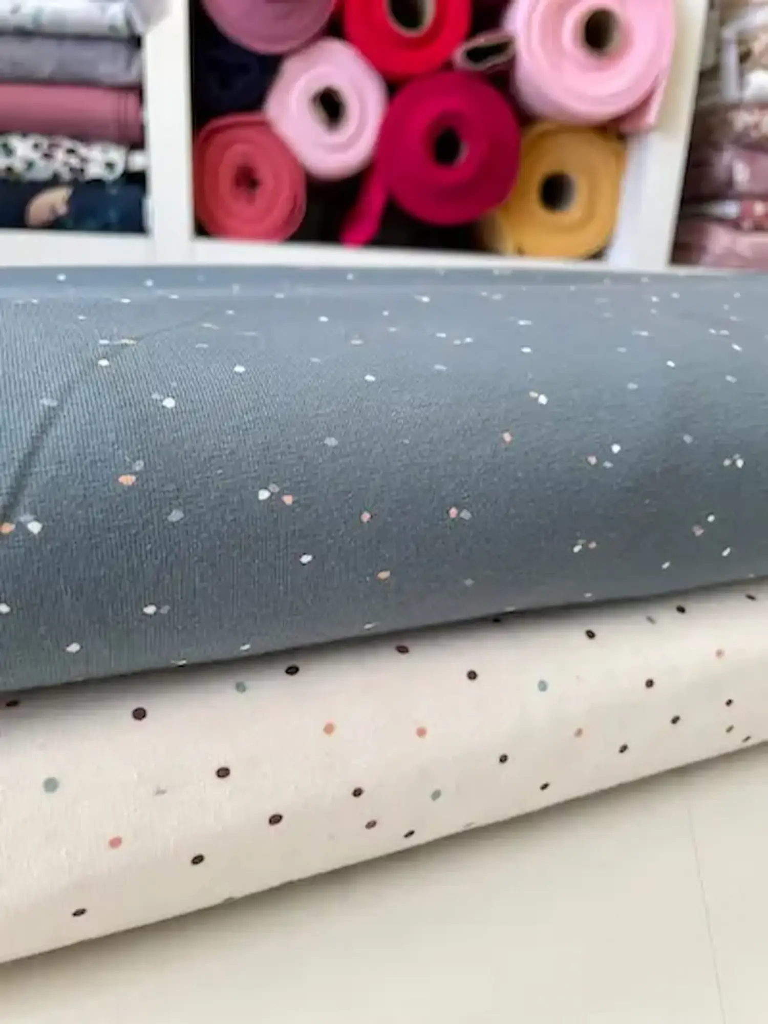 Zwei Rollen Baumwolljersey: oben blaugrau mit hellen Sprenkeln, unten wollweiß mit bunten Pünktchen.