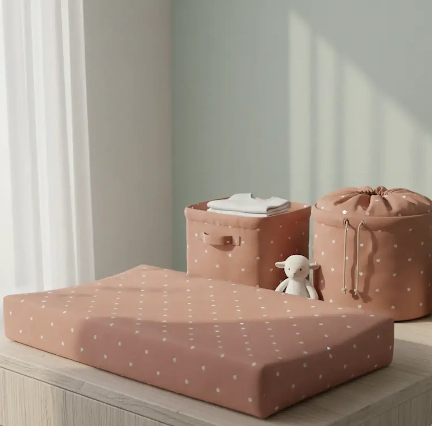 Babyzimmer-Zubehör aus robuster Baumwolle-Webware mit einem feinen, weißen Allover-Herzen-Muster auf terracottafarbenem Grund. Die Szene zeigt eine gemusterte Wickelauflage, eine quadratische Aufbewahrungskiste und einen passenden Wäschebeutel mit Kordelz