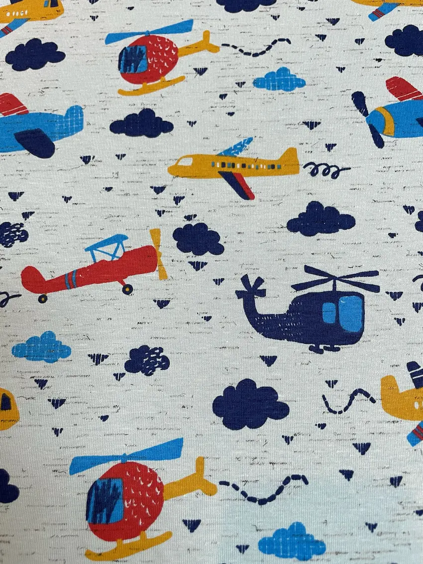 Ein flach liegender Baumwolljersey-Stoff als Meterware in Hellgrau mit Struktur-Optik, bedruckt mit einem farbenfrohen Kindermuster aus roten, blauen und gelben Flugzeugen, Hubschraubern und dunkelblauen Wolken.