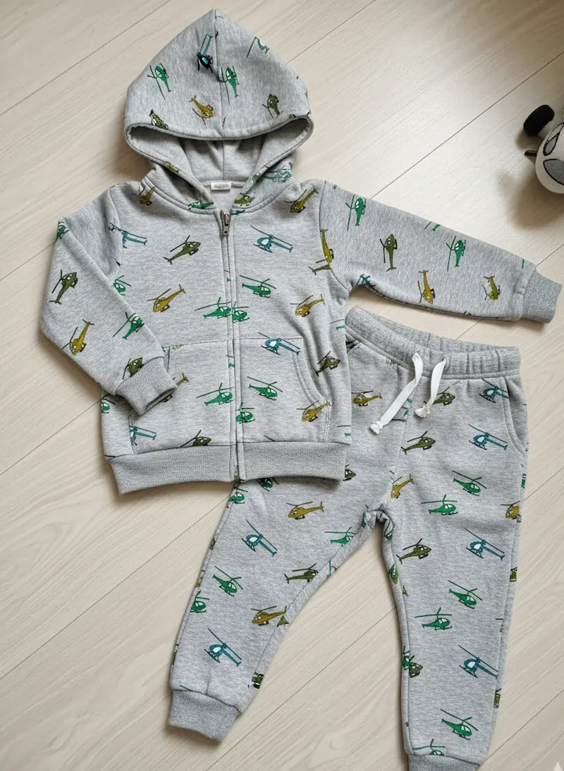 Ausgelegter Kinder-Sweatanzug aus hellgrau meliertem Alpenfleece mit Hubschrauber-Motiv, bestehend aus Hoodie mit Kapuze und passender Jogginghose.