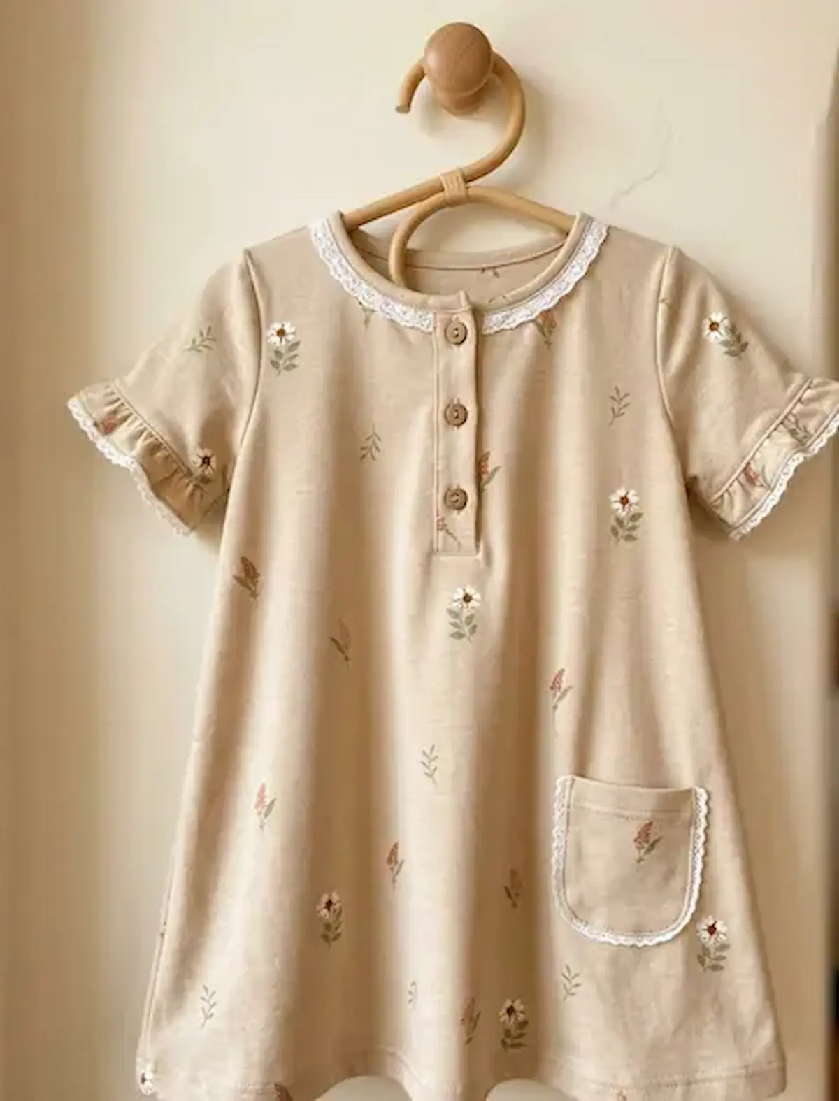 Handgenähtes Kinderkleid aus beigem Jersey mit Wildblumen-Print, weißer Klöppelspitze am Ausschnitt und Holzknöpfen auf einem Holzkleiderbügel.