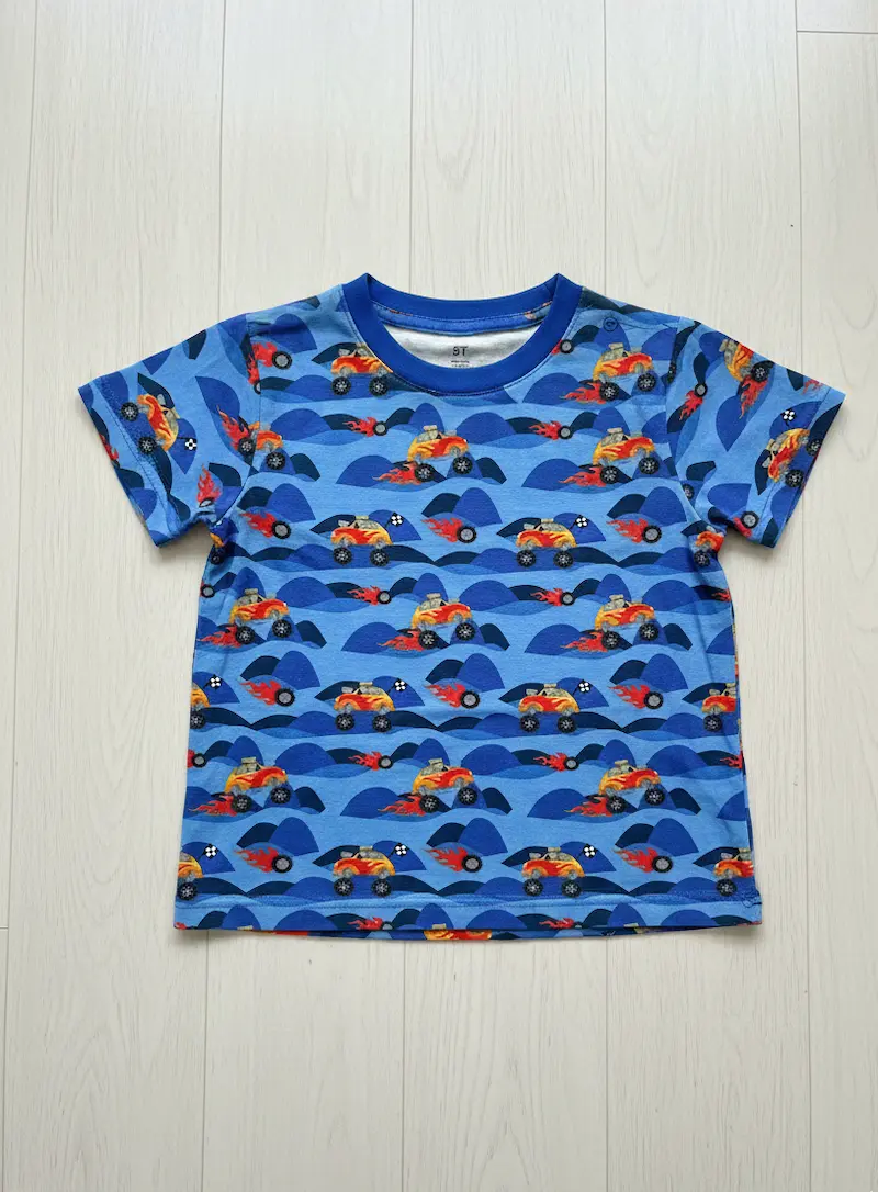 Eine flach liegende Kinderhose aus blauem Baumwolljersey mit einem Allover-Print von Monster-Trucks mit Flammenmotiven vor einer hügeligen blauen Landschaft. Die Hose ist mit passenden royalblauen Bündchen an den Beinen und einem elastischen Bund versehen