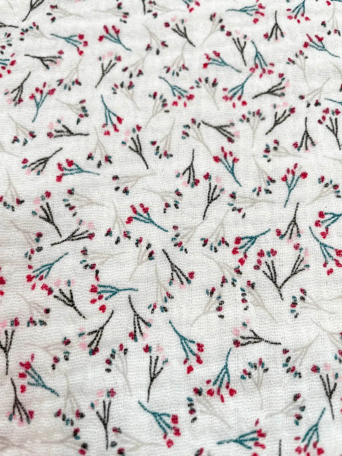 Flächenansicht von weißem Musselin-Stoff mit einem dichten, all-over Print aus filigranen Beeren-Zweigen in Rot und Blau. Zeigt die Musterwiederholung der Double-Gauze Meterware.