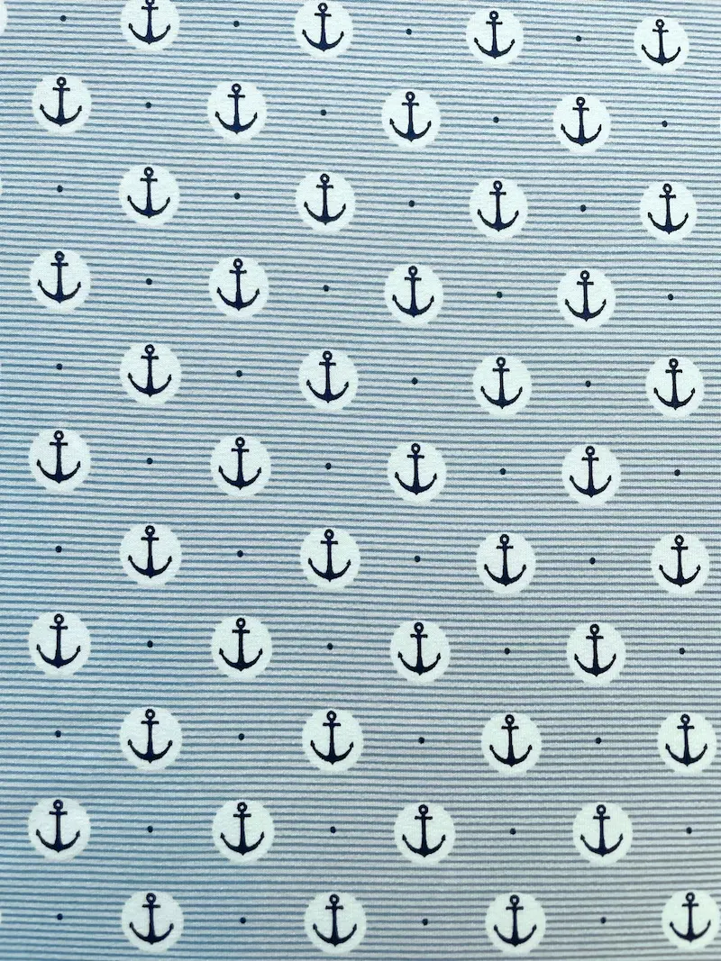 Nahaufnahme von elastischem Baumwolljersey in Blau-Grau feingestreift, bedruckt mit dunkelblauen Ankern in weißen Kreisen und kleinen Punkten; hochwertige maritime Meterware für Kinderkleidung und modische Accessoires.