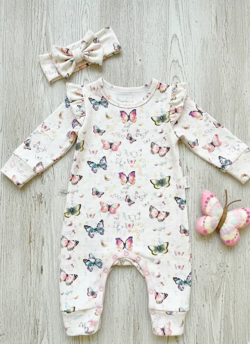 Ein cremefarbener Baby-Strampler aus Modal French Terry mit bunten Aquarell-Schmetterlingen. Das Kleidungsstück liegt flach auf hellem Holz und zeigt Details wie Schulterrüschen und farblich passende Accessoires (Stirnband und Stoffschmetterling).