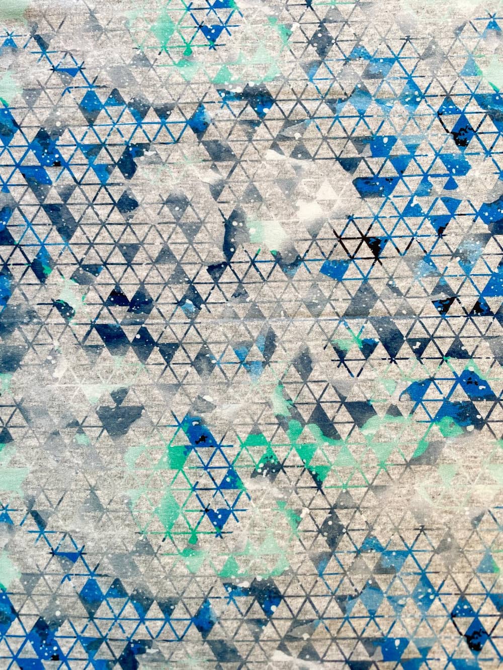 Nahaufnahme von French Terry Meterware mit einem modernen geometrischen Muster aus blauen und türkisfarbenen Dreiecken auf grau meliertem Hintergrund.