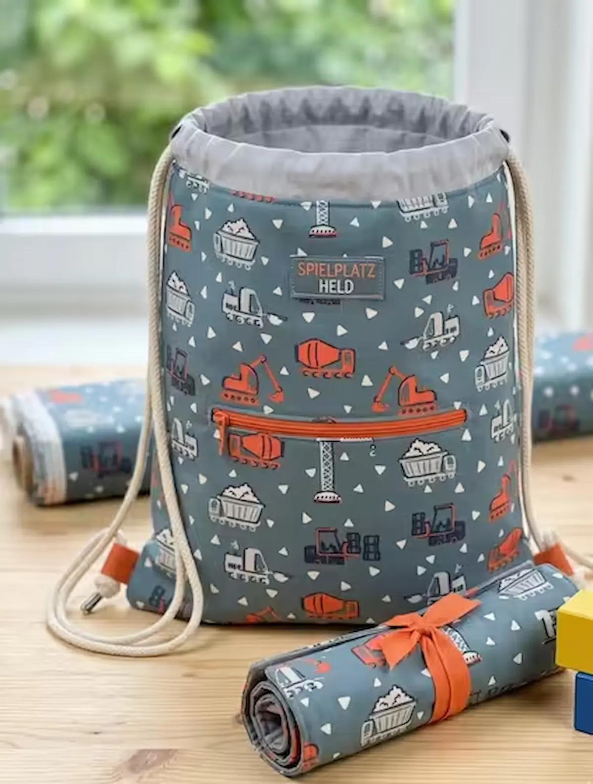 Hochwertiger Baumwollstoff als Meterware in Graublau mit Baustellen-Motiven wie Baggern, Kranen und Betonmischern, verarbeitet als genähter Rucksack mit der Aufschrift Spielplatz-Held.