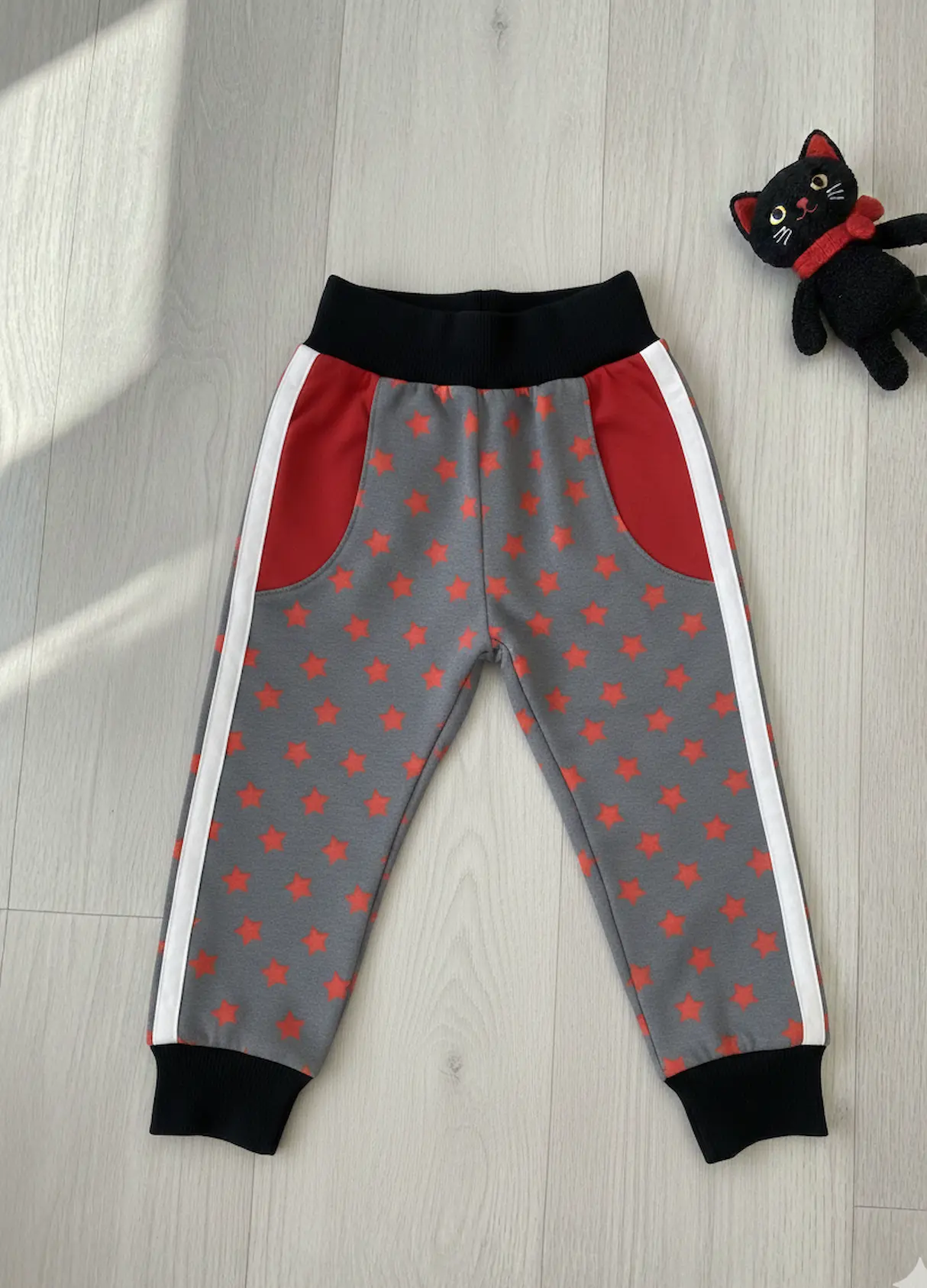 Nähbeispiel für grauen Sternen-Sweatstoff: Selbstgenähte Kinder-Jogginghose mit roten Taschenbelegen und schwarzen Bündchen auf hellem Holzboden