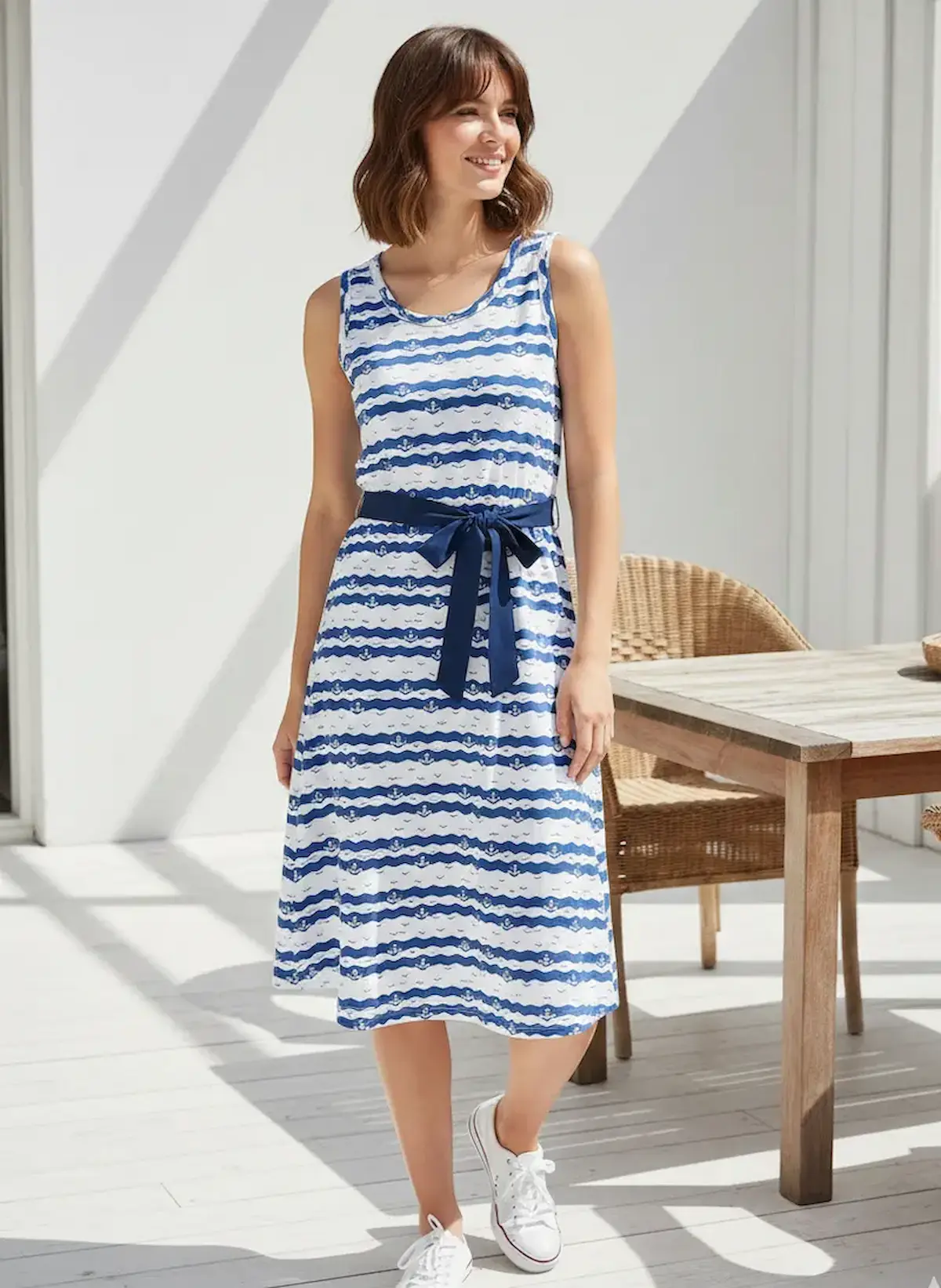 Eine lächelnde Frau in einem knielangen Sommerkleid mit Taillengürtel, genäht aus maritimen Baumwolljersey mit blau-weißem Anker- und Wellenmuster.